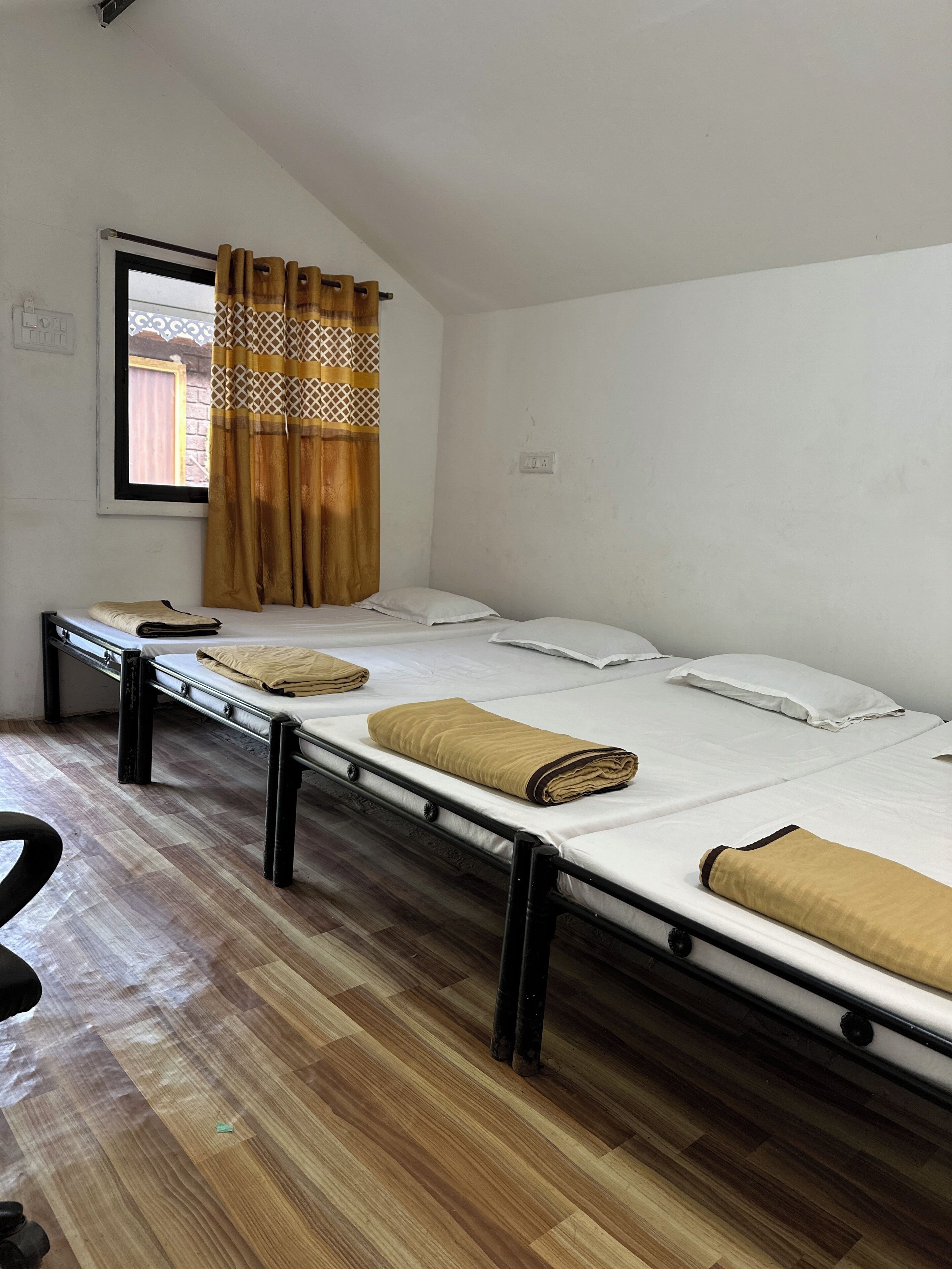 A dormitory room with two single beds, a window with patterned curtains, and a wooden floor.