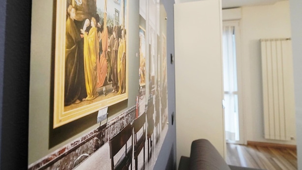 Un pasillo con un cuadro grande de estilo renacentista en la pared, un banco de mármol y vistas a una habitación con ventana y radiador.