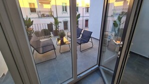 Eine Glastür führt zu einem Außenbalkon mit Sitzgelegenheit, Blumentöpfen und Blick auf benachbarte Gebäude.