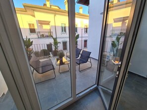 Puerta de cristal que da a un balcón exterior con zona de estar, plantas en macetas y vistas de edificios vecinos.