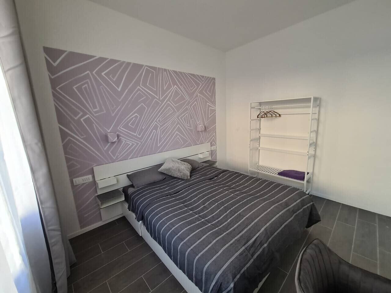 A bedroom with a bed, a white headboard, a patterned wall, a window with a white blind, and a small shelf.