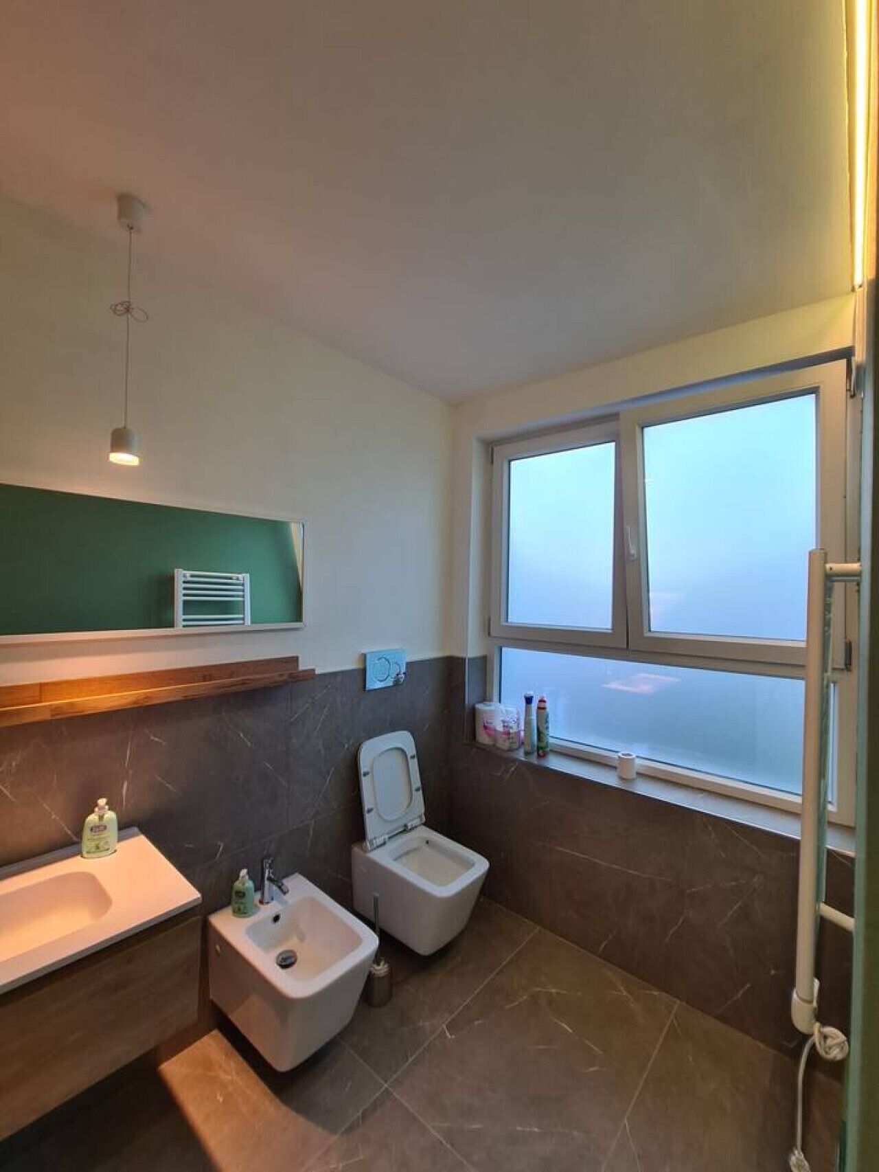 Un bagno moderno con WC, bidet e lavandino. Una finestra permette luce naturale e vista sul cielo.