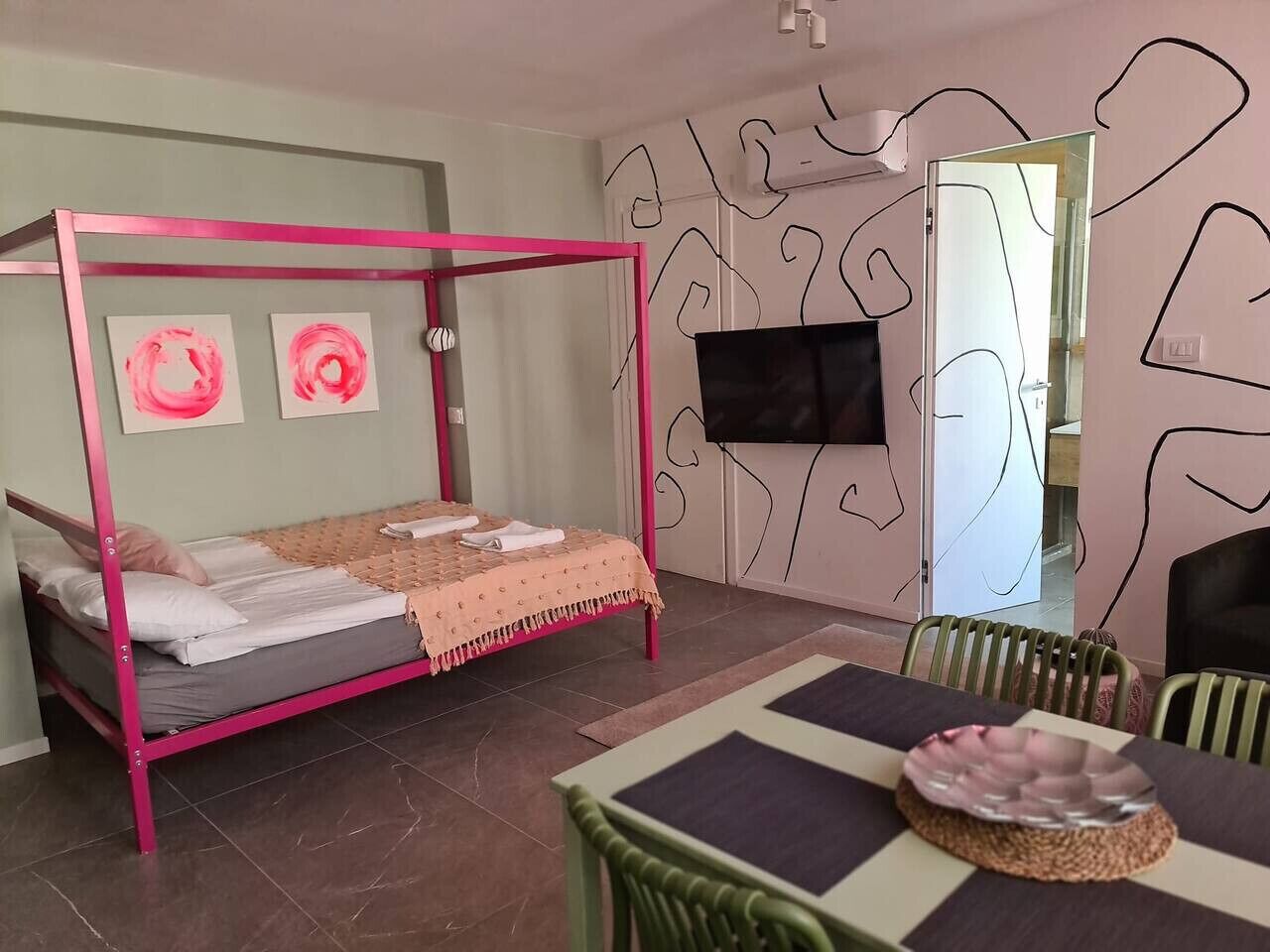 A room with a pink four-poster bed, a dining table with chairs, and abstract wall art.