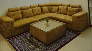 Un salon avec un grand canapé en forme de L, une table basse en verre, un tapis à motifs, un ventilateur de plafond et un tableau accroché au mur.