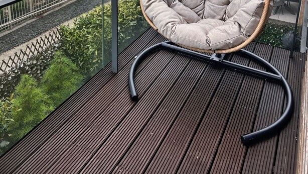A wooden deck with a wicker chair, a metal railing, and a view of a brick building with balconies.