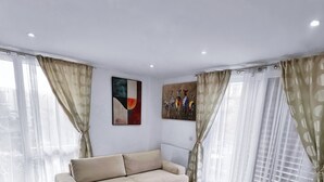 A modern living room with a beige sofa, a wooden coffee table, and a brown rug. There are two abstract paintings on the walls and sheer curtains on the windows.