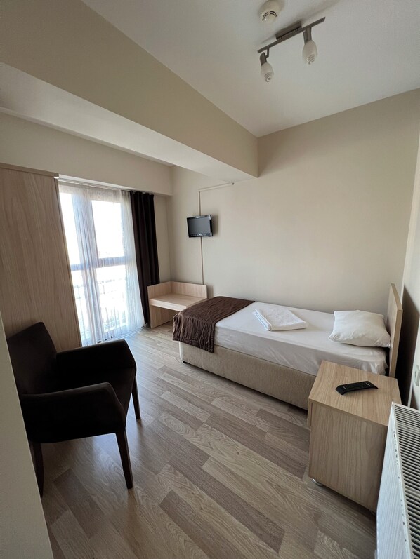 A hotel room with a bed, a desk, a chair, a TV mounted on the wall, and a window with curtains. - AKAR PANSİYON (Çanakkale)