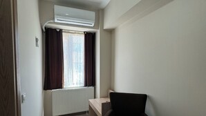 A small, well-lit room with a wooden floor, a black chair, a small desk, a radiator, and a window with curtains.