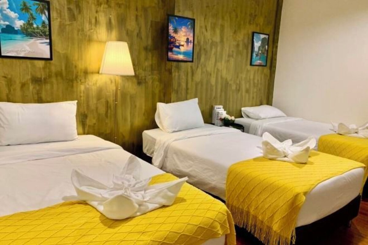 A hotel room with three beds, each with white linens and a yellow quilted bedspread. There are wall-mounted lamps and framed pictures depicting tropical scenes.