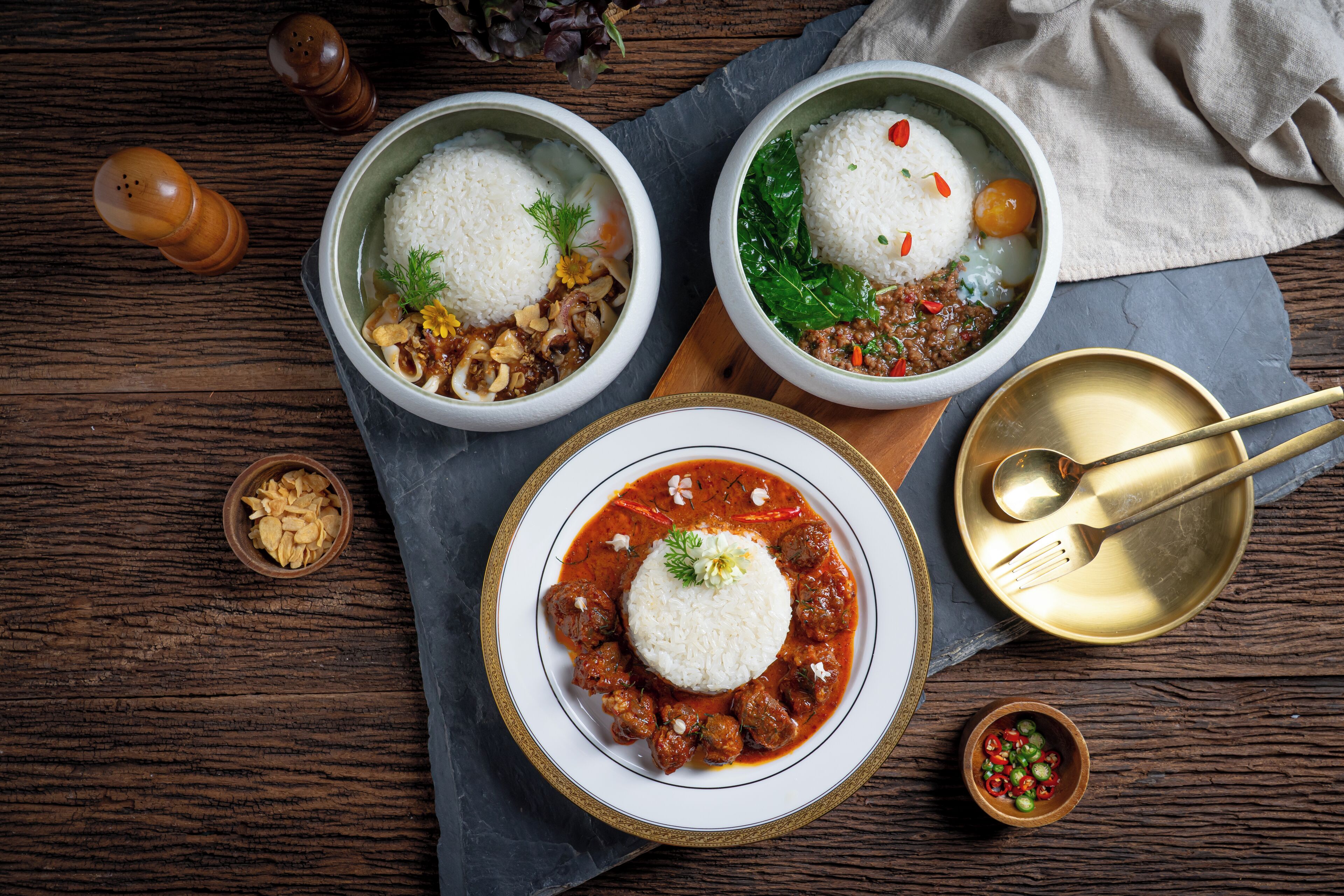 Three bowls of rice with different toppings, a bowl of chili, and a bowl of sliced peppers on a wooden table.