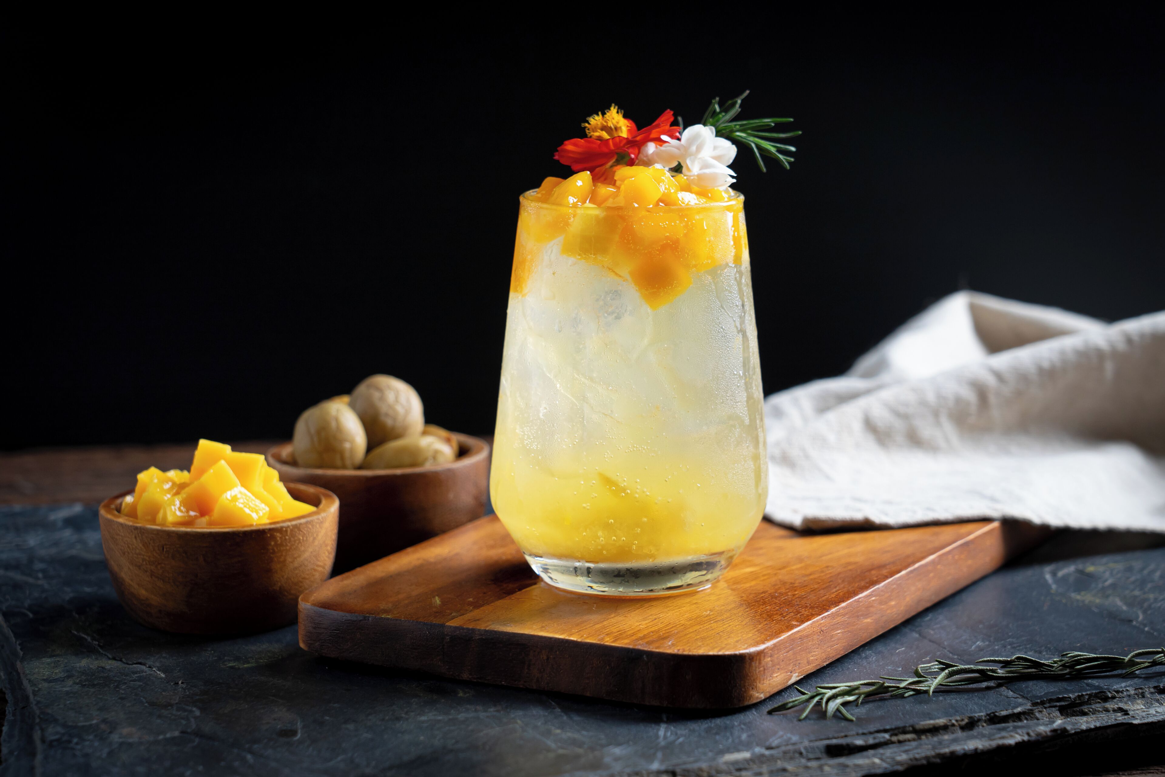 Un vaso alto con una bebida en capas, decorado con frutas y flores, sobre una tabla de madera con fondo oscuro.