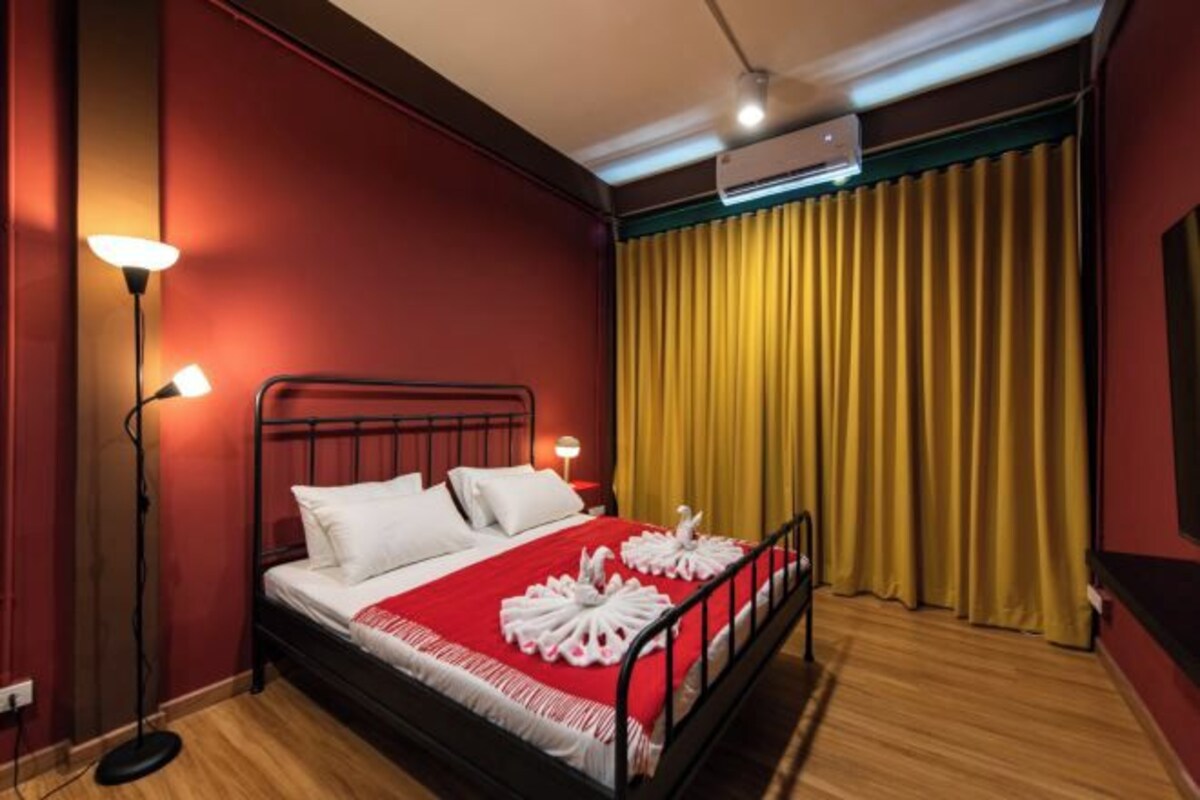 A hotel room with a red wall, a black metal bed frame, a red and white bedspread, two white pillows, a bedside table with a lamp, and a yellow curtain.