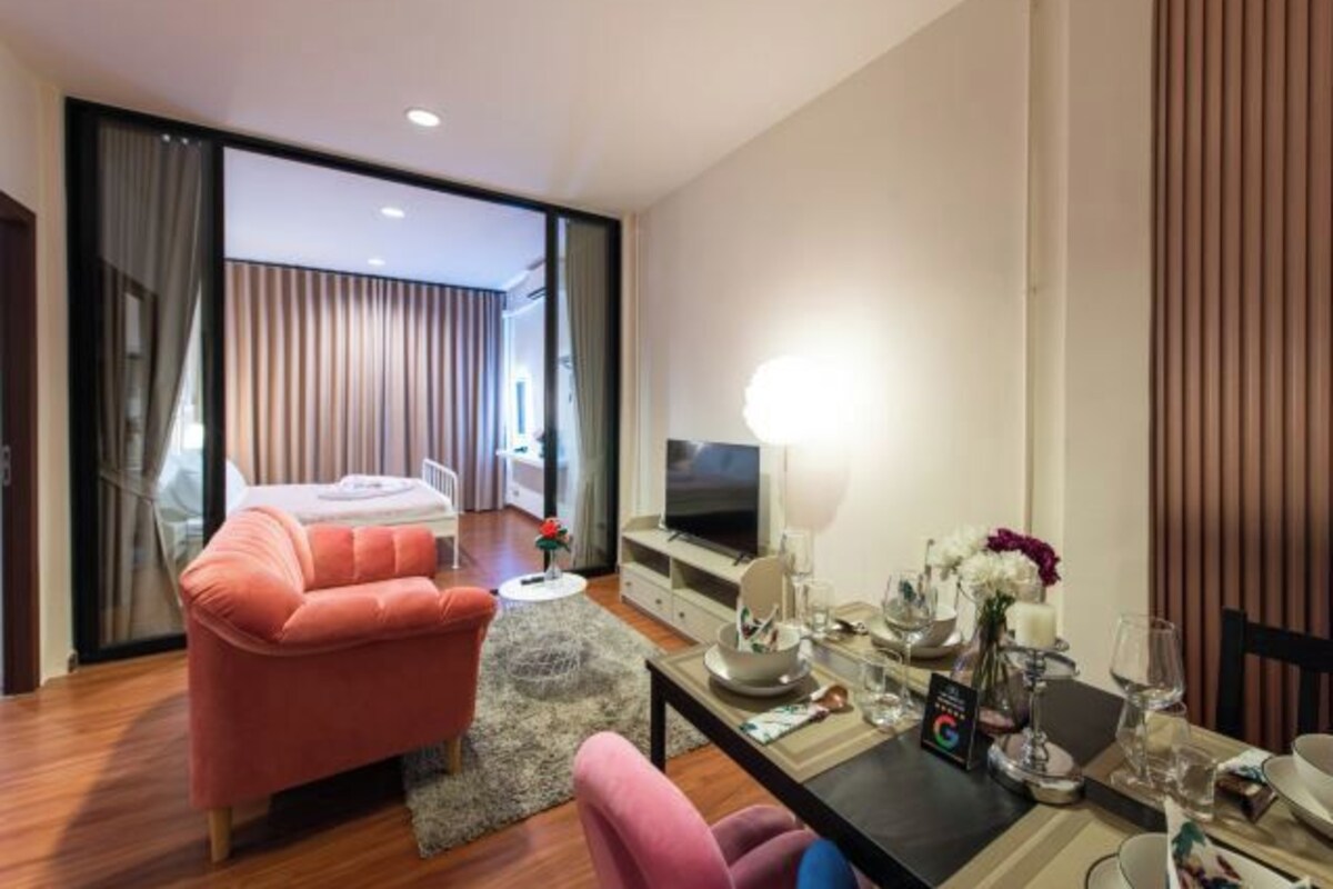 A modern hotel room with a sofa, a dining table set for two, a TV, and a large window with curtains.