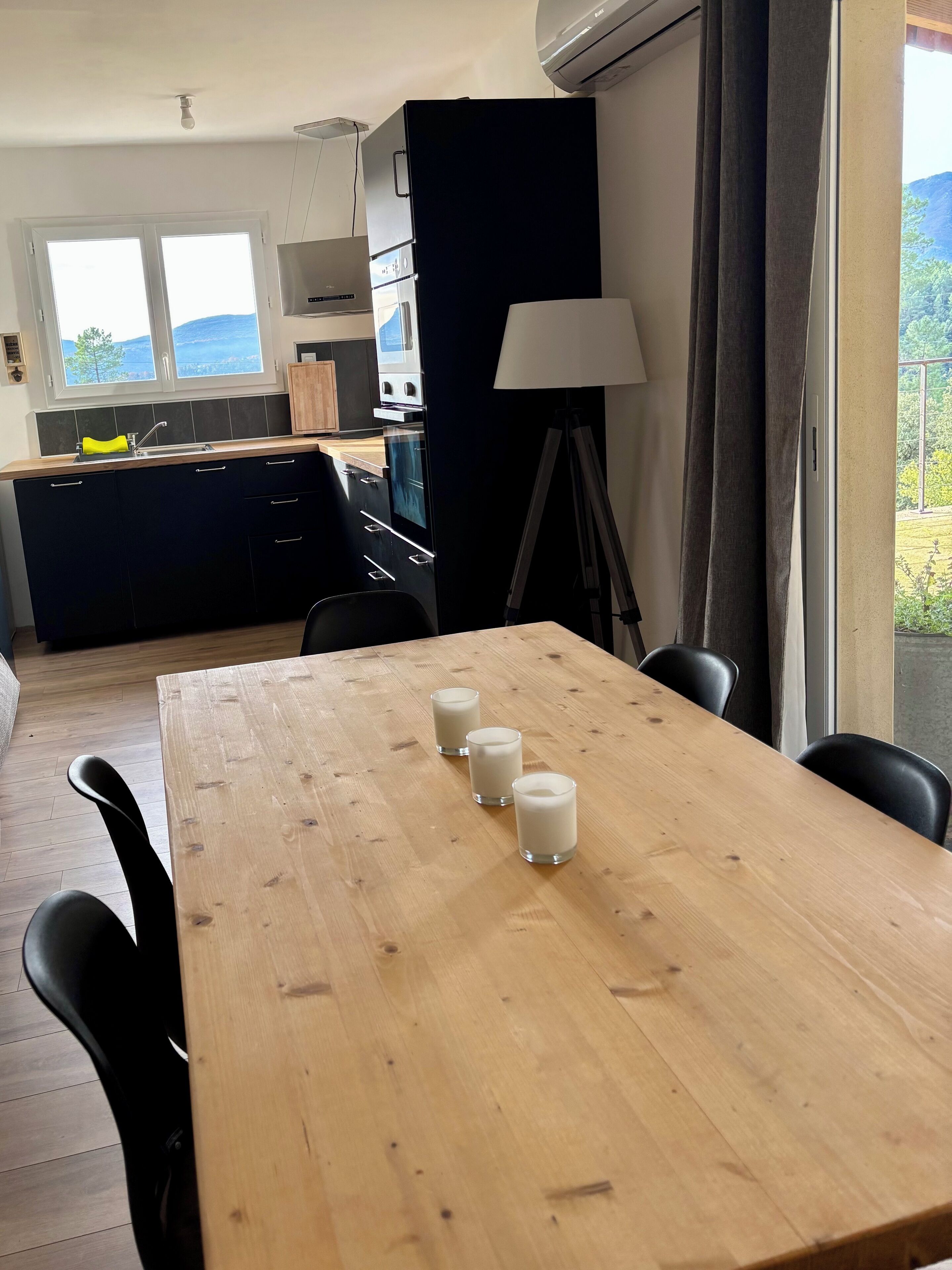 A modern kitchen with a wooden table and chairs, a dining area with a lamp, and a view of the outdoors through a window.