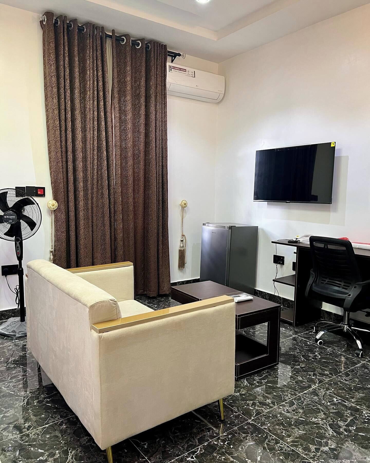 A hotel room with a sofa, a small table, a TV, a fan, and a minibar.