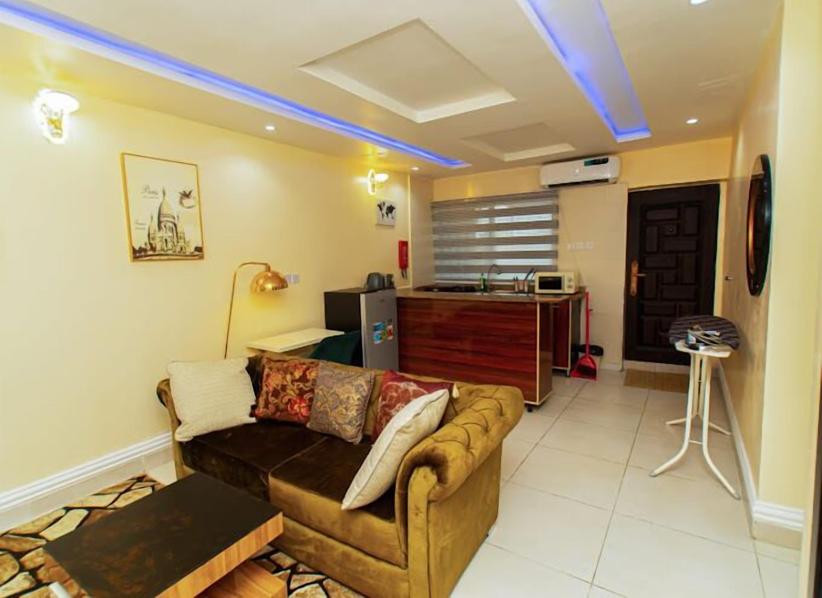 Uma sala de estar com sofá, mesa de centro, luminária e um espaço com cozinha compacta, incluindo micro-ondas e geladeira.