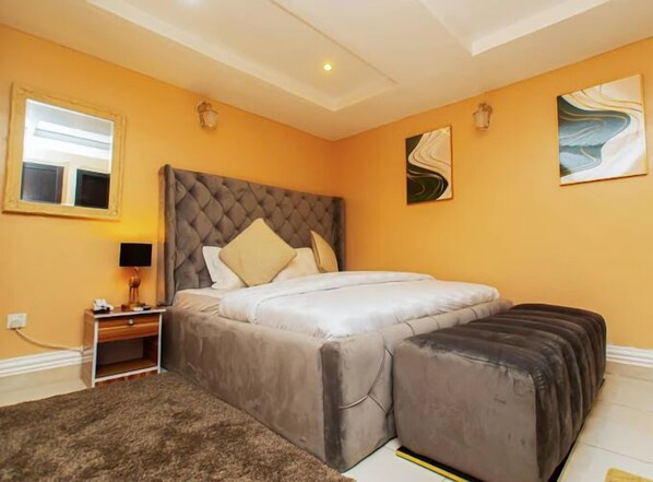 A bedroom with a large bed, a bedside table with a lamp, and a small desk. - His Glory's Apartment (Abuja)