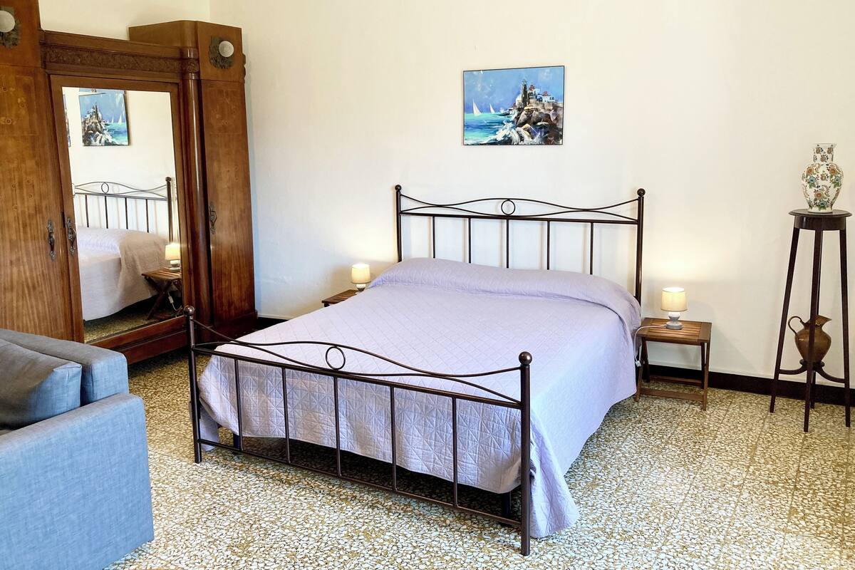 Een slaapkamer met een bed, een houten kledingkast, een kleine tafel met een lamp, een decoratieve vaas en een schilderij aan de muur.