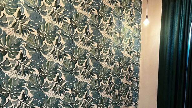 A neatly made bed with a white comforter and blue pillows, a bedside table with a lamp, and a wall with a tropical leaf pattern. - Perfect for exploring Naples without having to use transportation.  (Napoli)