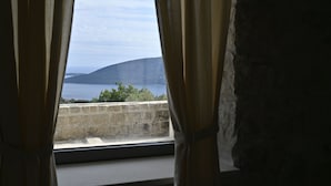 A view through a window with curtains, featuring a stone wall and a distant view of a hill and water body. - Dreamlike stone house by the sea: luxury with infinity pool and panoramic views (Herceg Novi)