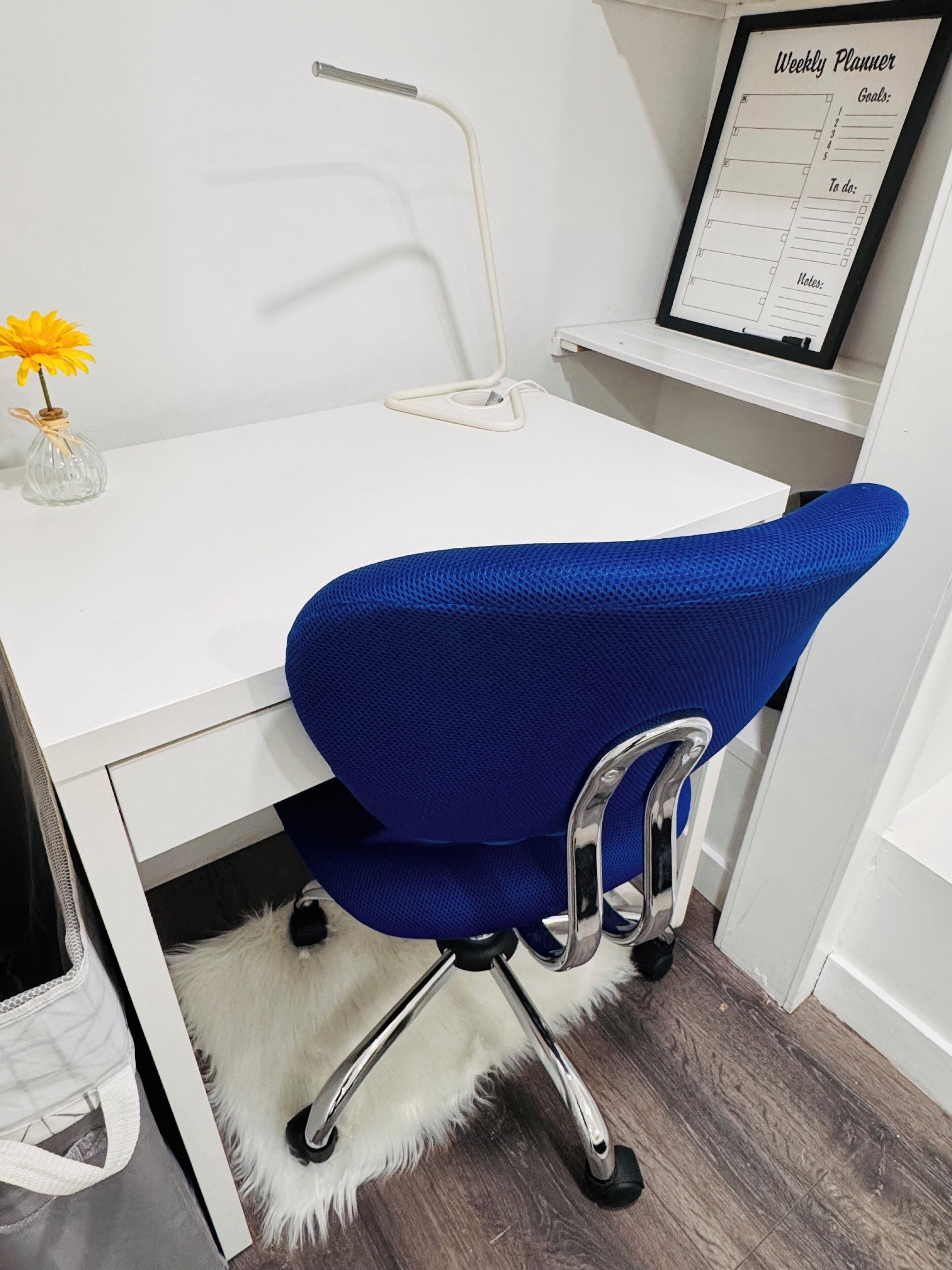 A white desk with a blue office chair, a framed weekly planner, and a small yellow flower vase.