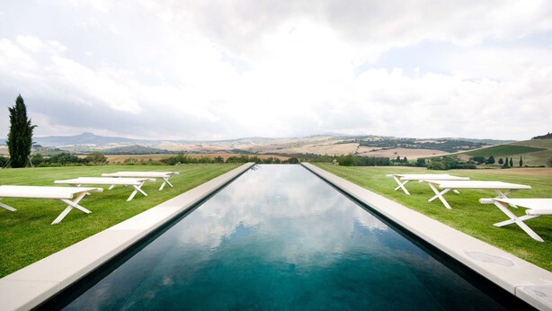A swimming pool with white lounge chairs on either side, surrounded by a grassy area with a view of distant hills. - Villa Olimpia in Sarteano (Sarteano)