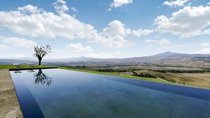 Piscina límpida com uma árvore no centro, rodeada por uma área gramada e vista para colinas ao longe.