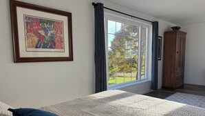 A bedroom with a large window, a bed with a quilted cover, a wooden wardrobe, and a framed picture on the wall. - Beautiful new studio with amazing views - peaceful but close to everything! (Weston)