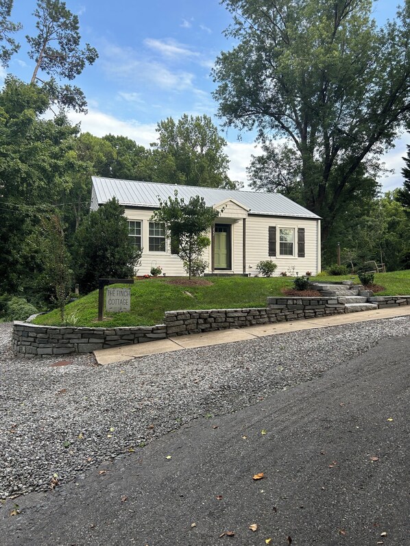 A single-story cottage with a metal roof, surrounded by a stone retaining wall and a gravel driveway.