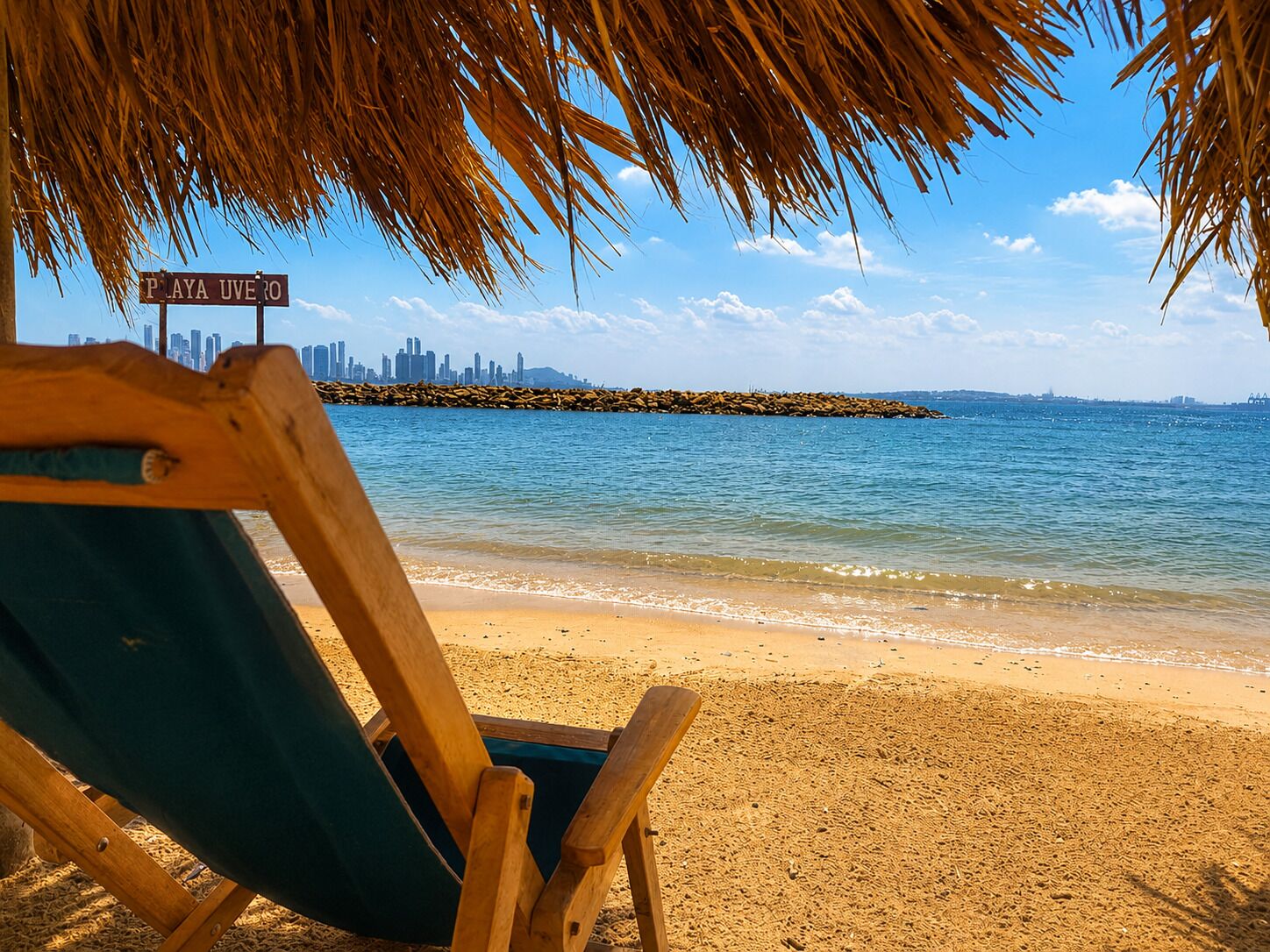 A beach chair under a palm tree with a city skyline in the background.
