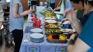 A communal dining area with a long table covered by a patterned tablecloth, various food items, and people engaged in conversation.
