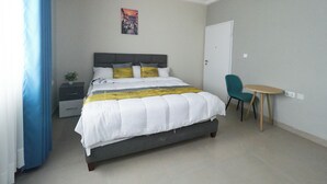 Quarto de hotel bem arrumado com cama, uma mesinha, uma cadeira, um criado-mudo e um aparelho de ar condicionado.