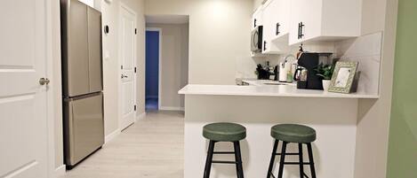 Ett modernt kök med vita skåp, ett kylskåp i rostfritt stål och en liten bardisk med barstolar.