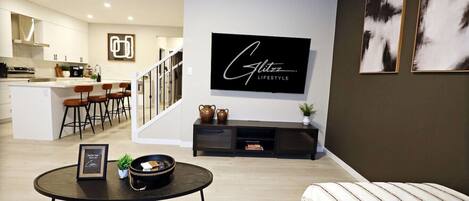 A modern living room with a black-framed TV, a round coffee table, and a sofa with a striped throw. A staircase leads to a kitchen area with a bar and seating.