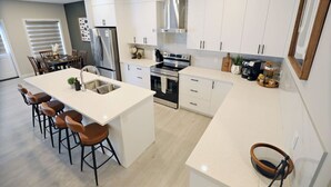 Une cuisine moderne avec des meubles blancs, un îlot central doté de tabourets, des appareils en acier inoxydable et un coin repas avec une table et des chaises.