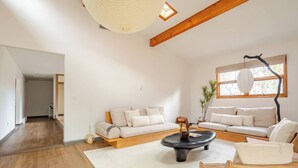 Un salon spacieux avec un plafond haut, des poutres apparentes, une grande fenêtre offrant une vue dégagée, un ensemble de canapés, une table basse et une table d’appoint.