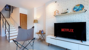 Ein modernes Wohnzimmer mit weißer Backsteinwand, Flachbildfernseher, grauem Sessel, Holztür und einer Treppe.