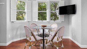 A dining area with a round table and six wicker chairs, three windows with white shutters, and a flat-screen TV mounted on the wall. - Waverly Suite 2 (New Shoreham)