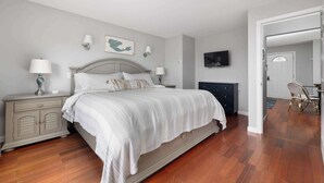 A bedroom with a large bed, a nightstand with a lamp, a dresser, and a television on the wall. - Waverly Suite 2 (New Shoreham)