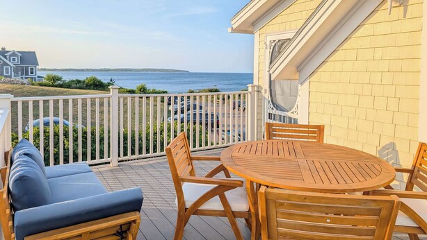 A wooden deck with a round table and chairs, overlooking a body of water and a distant shoreline. - Waverly Suite 3 (New Shoreham)