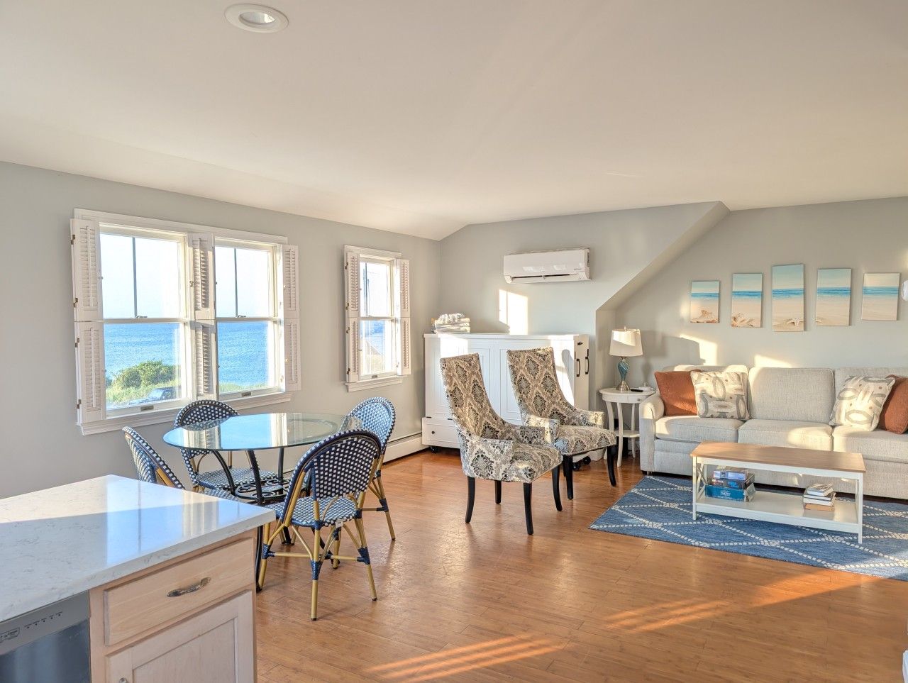 A spacious living area with a dining table, chairs, a sofa, and a coffee table. There are windows with shutters, an air conditioning unit, and artwork on the walls.