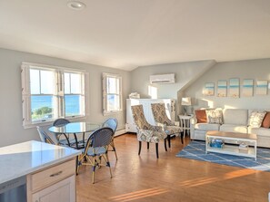 Un vaste espace de vie comprenant une table à manger, des chaises, un canapé et une table basse. On y trouve des fenêtres avec volets, un appareil de climatisation et des œuvres d’art accrochées aux murs.