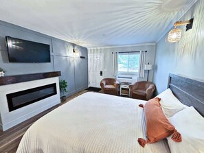 Une chambre d’hôtel dotée d’un grand lit, d’un téléviseur à écran plat, d’une cheminée, de deux fauteuils, d’une petite table et d’une fenêtre avec des rideaux.