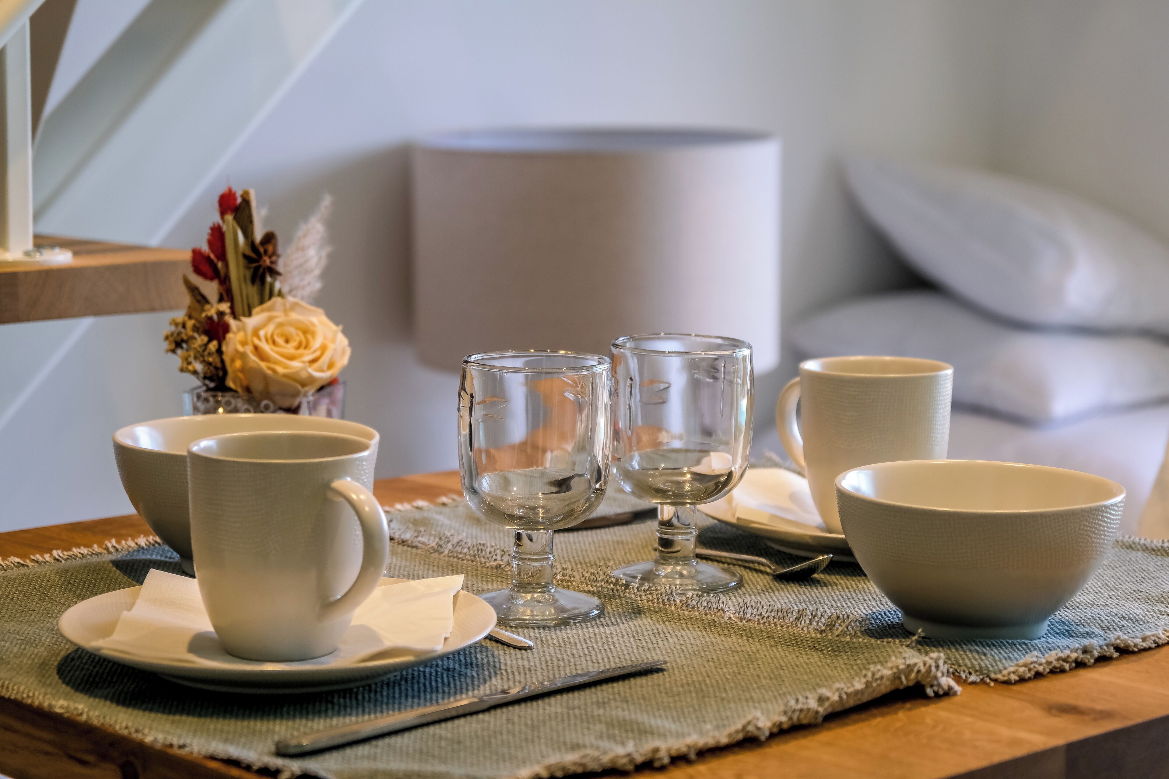 Una mesa de comedor dispuesta con tazas, cuencos y vasos de cerámica blanca sobre un mantel tejido.