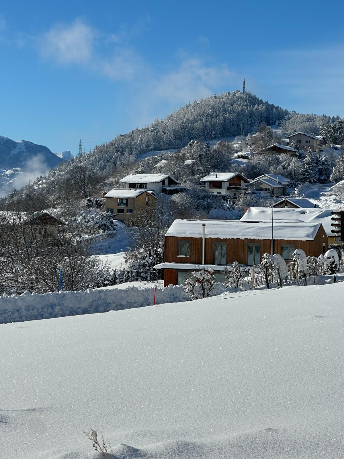 Un quartier résidentiel enneigé, avec des maisons construites sur une colline, un ciel dégagé et une montagne visible au loin.