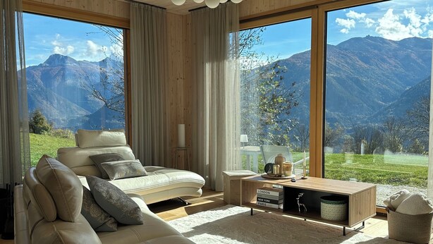 Un salón moderno con un sofá grande, una mesa de centro y vistas a montañas a través de amplios ventanales.