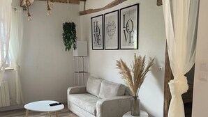 Sala de estar com teto de madeira, sofá, mesa pequena e abajur. Há quadros nas paredes e uma planta ao lado.