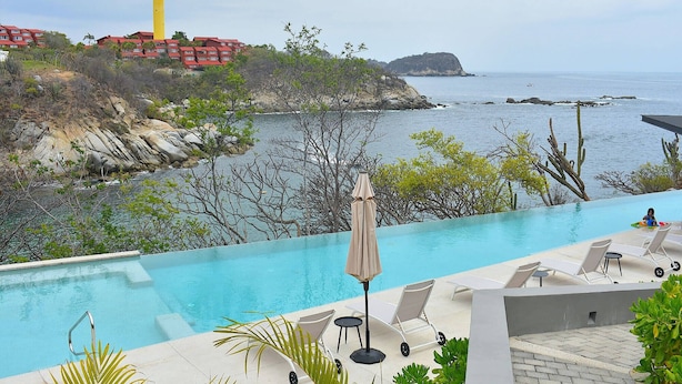 Un espace aménagé au bord de la piscine, avec des chaises longues et un parasol, offrant une vue sur un paysage côtier avec des bâtiments et une côte rocheuse.