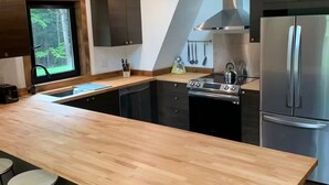 A modern kitchen with a wooden island, bar stools, stainless steel appliances, and a view of the forest through a window.