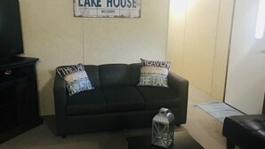 Una sala de estar con un sofá oscuro, dos almohadas, una mesita con un recipiente de vidrio y un letrero en la pared que dice 'LAKE HOUSE'.